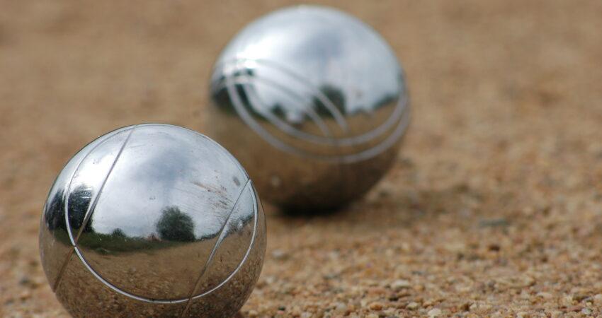 Zwei silberglänzende Boule-Kugeln liegen im Sand. In ihnen spiegeln sich unscharf der Himmel und ein paar Bäume.
